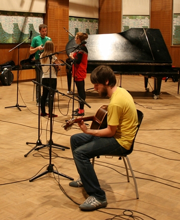© radio fm4 gstettner gitarrist von seabear sitzt auf einem stuhl und spielt gitarre, davor mikros, dahinter ein fluegel und andere bandmitglieder