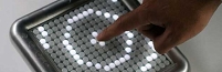 © oddstrument.com Das elektronische Musikinstrument Tenori on, auf das mit dem Finger gezeigt wird.