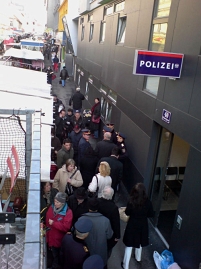 Menschen zwischen einer Häuserfassade mit Polizeischild und der Rückseite von Marktständen am Brunnenmarkt in Wien-Ottakring