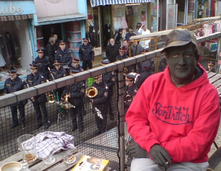 ein schwarz geschminkter Mann im roten Sweater sitzt au einem Balkon ein Stockwerk über dem Brunnenmarkt, wo eine Polizeikapelle spielt