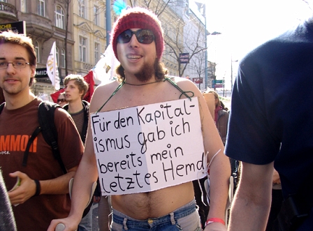 Demonstration in Wien; junger Mann mit Mütze und nacktem Oberkörper hat Schild umgehängt, auf dem "Für den Kapitalismus gab ich mein letztes Hemd" steht