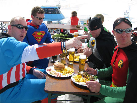 vier Männer als Superhelden verkleidet essen Pommes und trinken Bier vor einer Schihütte.