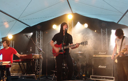 Ein Sänger mit Kapuzenpulli singt energisch ins Mikro. Links ein konzentrierter Keyboarder, rechts ein in sich versunkener Bassist.