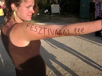 eine frau hat sich auf ungarisch "festival" auf den arm geschrieben und hält den schriftzug in die kamera. im hintergrund: sonnenuntergang