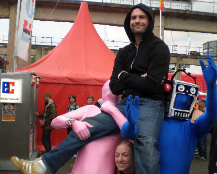 zwei Festivalbesucher in Lycra-Anzügen stemmen einen dritten im Kapuzenpulli auf ihren Schultern in die Höhe.