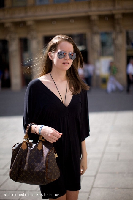 © Daniel Troyse Mädchen mit Sonnenbrille in kurzem schwarzem Kleid mit Vuitton Logo Canvas Tasche über dem linken Arm.