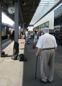 Älterer Herr steht auf Bahnsteig und schaut. Entfernt steht Menschenansammlung.