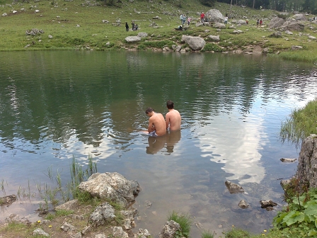 Am Bergsee. Am Ufer einige Nonnen und Wanderer. Im Wasser sitzen zwei Menschen.