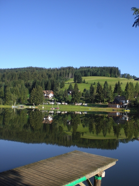 Ein kleiner See, im Vordergrund ein Steg. Im ausnahmslos glatten Wasser eine perfekte Spiegelung des gegenüberliegenden Ufers.