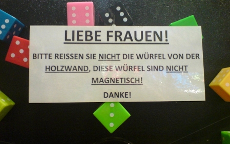 Schild mit Aufschrift: Liebe Frauen. Bitte reißen sie nicht die Würfel von der Holzwand, diese Würfel sind nicht magnetisch