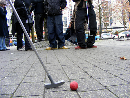 Ein Minigolf-Schläger mit Ball, im Hintergrund drei Menschen, die mit ihren Füßen eine Barriere bilden.