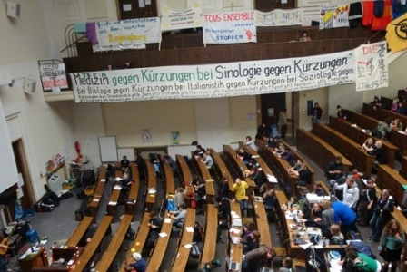 Ein Hörsaal. In den Bänken sitzen einige Studenten, an den Ballustraden sind Transparente angebracht.