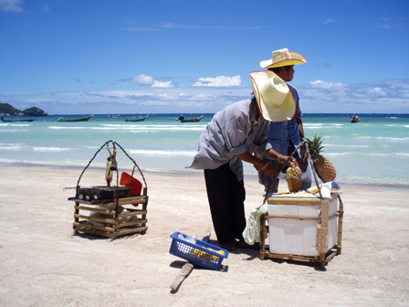 Thailand - heller Strand, tuerkises Meer, blauer Himmel, zwei Maenner mit Strohhueten, die Ananas aufschneiden und verkaufen.