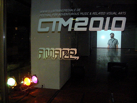 Durchsichtige Glasscheibe mit darauf geklebten Logos von der club transmediale und A MAZE.-Festival. Im Hintergrund ist ein großer Screen zu sehen.