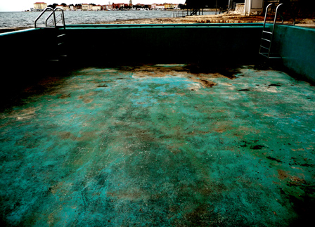 © andrea neustifter leerer swimmingpool, der gruenlich rostet, im hintergrund das meer u ein dorf