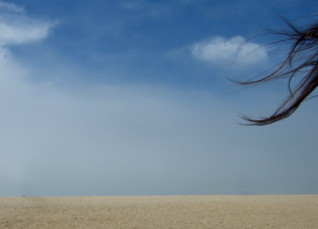 sandstrand und wolkenhimmel am rechten oberen eck flattern lange dunkle haare ins bild