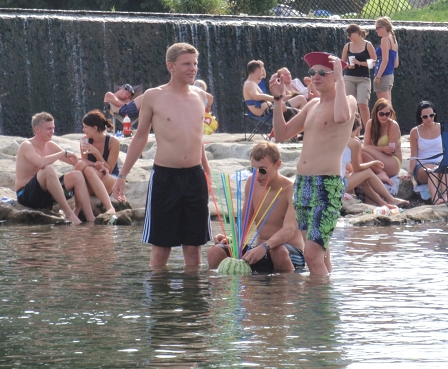 Burschen vor dem Wehr-Wasserfall trinken einen Cocktail aus einer schwimmenden Wassermelone.