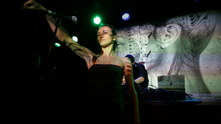 Tania Saedi mit ausgestrecktem Arm im grün-blauen Bühnenlicht. Hinter ihr das Albumcover an die Wand projiziert und Markus Kienzl am Mischpult.