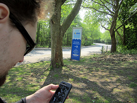 Ländliche Gegend bei Sonnenschein und ein Schild zum Strandbad. Im Vordergrund ein Mann, der auf sein Smartphone schaut.