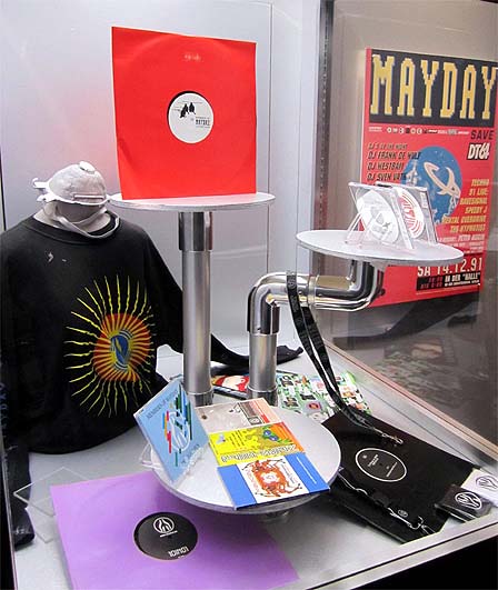 Platten, CDs und ein Shirt, hinter Glas aufbereitet. Dahinter ein Plakat der ersten "Mayday" aus dem Jahr 1991.