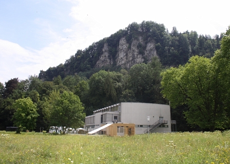 Die Blumenwiese, darin das alte Hallenbad, dahinter Wald und Felsen.