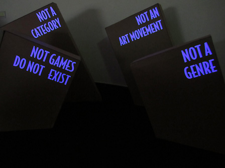 Vier Objekte aus Pappkarton, auf denen die Slogans "Not a category, "Notgames do not exist", "Not an art movement" und "Not a genre" geschrieben stehen.