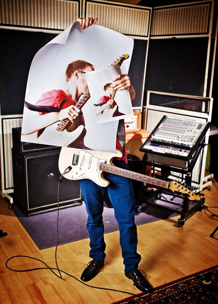 PeterLicht steht mit umgehängter Gitarre vor dem Verstärker und hält sich ein Poster über sein Gesicht.