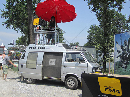 Ein silberner, älterer VW-Bus, der als DJ-Bus firmiert, darauf steht DJ JoeJoe mit Plattentellern und einem aufgespannten roten Sonnenschirm.