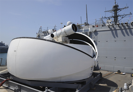 © US Navy Eine weiße, nach zwei Seiten geöffnete, halbkreisförmige Basisstation einer Laserkanone mit der Teleskop-förmigen Waffe in der Mitte.