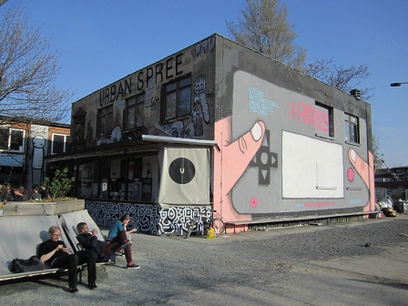 Vor dem Berliner Veranstaltungszentrum "Urban Spree": zwei Männer sitzen auf einer Bank, daneben ist eine große bemalte Wand, die einen Computerspiel-Controller darstellt.