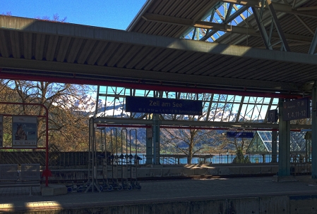 Ein Bahnsteig im Bahnhof von Zell am See, im Hintergrund der See und Schnee bedeckte Berge.