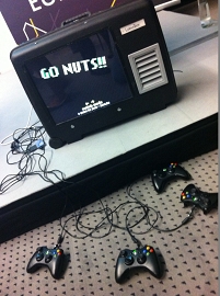 Ein Koffer, in dem ein Bildschirm eingebaut ist. Darauf dargestellt ist ein Schriftzug, der "Go Nuts!" anzeigt. Davor liegen einige schwarze Gamecontroller.