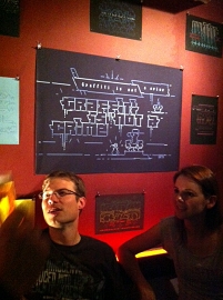 Zwei Besucher in der Bar, darüber und dahinter hängen Prints von ASCII-Art. Im größten Bild steht geschrieben: "Graffiti is not a crime".