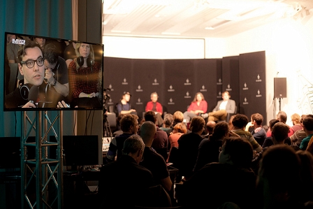 Gäste bei einer Paneldiskussion beim Elevate Festival 2013. Im Vordergrund ein Fernseher, auf dem der Hacker Jacob Appelbaum zu sehen ist.