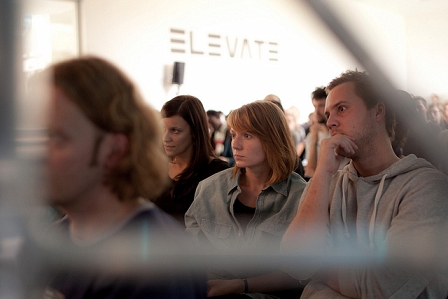 Zuhörerinnen und Zuhörer beim Diskursprogramm des Elevate Festivals. Dahinter ein großes Logo des Festivals in schwarz auf einer weißen Wand.