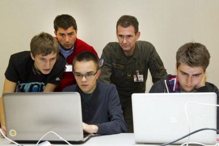 Junge Männer sitzen vor ihrem Laptop, zwei Herren schauen über ihre Schultern