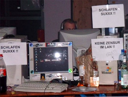 Computer und Bildschirme bei einer LAN-Party im Jahr 2000. Dazu ausgedruckte Zettel, auf denen "SCHLAFEN SUXXX!!" und "KEINE ZENSUR IM LAN!!" steht.