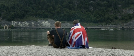 Zwei Buben sitzen am Ufer eines Sees, der eine mit Lederhose, der andere in eine britische Flagge eingehüllt.