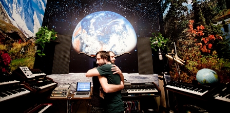 Die zwei Musiker von Le Tam Tam umarmen sich inmitten ihres Synthesizerparks, an den Wänden Fototapeten vom Planeten Erde und Landschaften