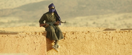 Ein vermummter Mann sitzt mit einem Gewehr auf einer Mauer
