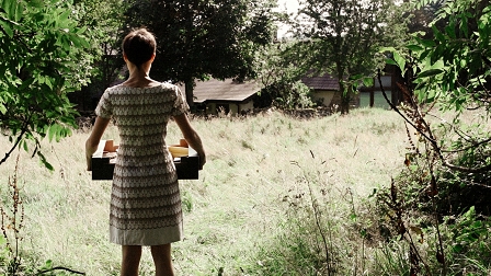 Eine Frau trägt einen Karton und steht im Garten eines Landhauses. Filmstill aus "Hide And Seek"