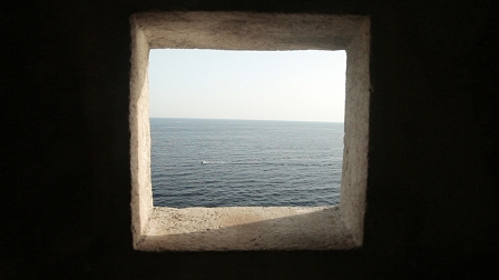 Blick durch ein Fenster in einer steinernen Hafenmauer auf das offene Meer