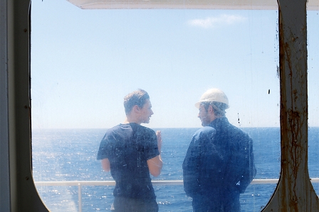 Vor einer Glasluke stehen zwei junge Männer an Bord eines Frachtschiffes und schauen auf das Meer. Filmstill aus "Lichtes Meer"