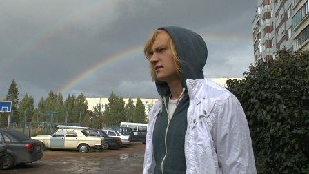 Jugendlicher mit langen blondierten Haaren trägt einen Kapuzensweater und geht über einen Parkplatz, im Himmel ein Regenbogen