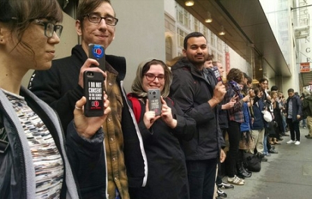 Menschen in einer Schlange halten iPhones mit einem Aufkleber "I do not consent" in die Höhe