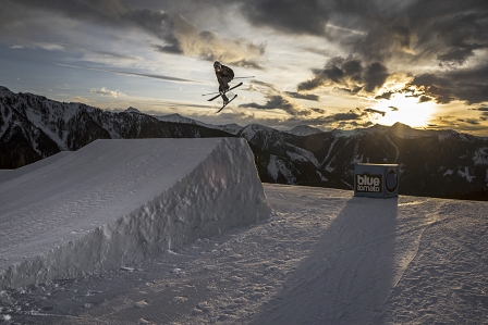 Skifahrerin springt über einen großen Kicker, im Hintergrund sieht man Berge und den Sonnenuntergang