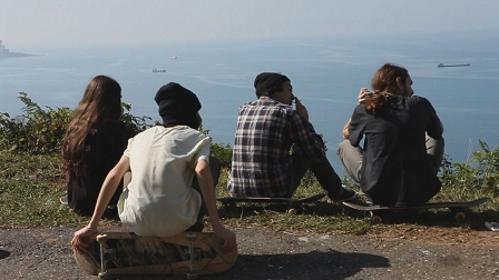 Vier männliche Jugendliche sitzen mit Skateboards am Straßenrand und schauen auf das Meer. Filmstill aus "When the Earth seems to Be Light"