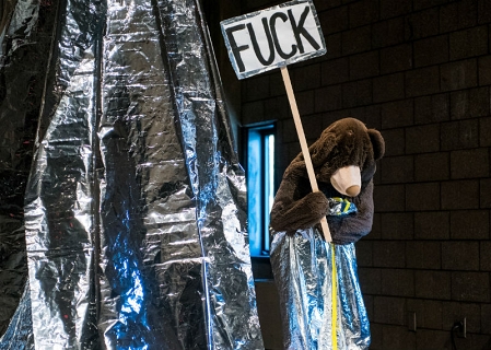 Ein großer Plüschbär auf einer Bühne. Er hält ein Schild mit der Aufschrift "Fuck".